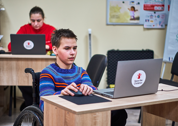 Ein Kind sitzt am Schreibtisch und blickt auf einen aufgeklappten Laptop mit Save-the-Children-Logo.