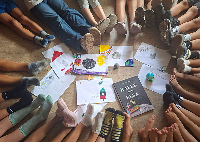 Kinder sitzen um ein Buch © Save the Children Eine Gruppe Kinder sitzt mit ausgestreckten Beinen um eine Sammlung von selbst gemalten Bildern und ein Kinderbuch herum