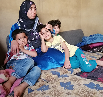 Eine Kopftuch-tragende Frau sitzt mit drei Kindern auf Kissen am Boden