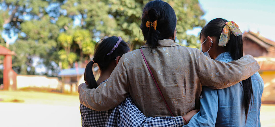 Eine Mutter umarmt ihre beiden Töchter. Die Familie musste nach der Eskalation der Gewalt vor einem Jahr aus ihrem Dorf fliehen. Nun leben sie in einem Camp für Binnenvertriebene in Myanmar. © Save The Children