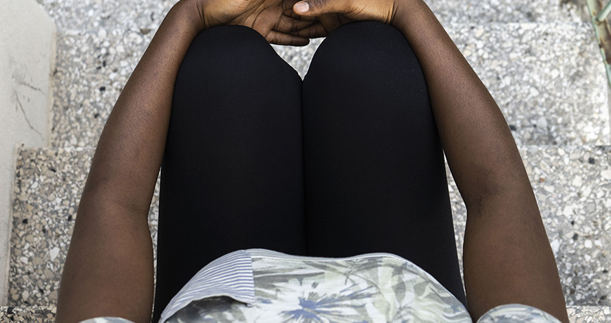 Oberkörper und Beine einer schwarzen Frau sind zu sehen, die die Arme um die Knie gelegt hat und auf einer Treppe sitzt. © Sara Mignogna/ Save the Children