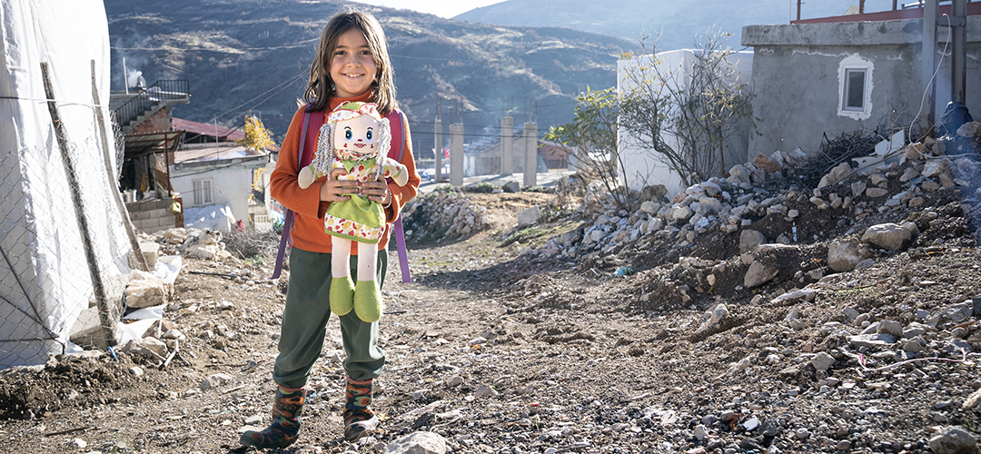 Ein Mädchen hält eine große Stoffpuppe im Arm und steht vor durch das Erdbeben zerstörten Häusern in der Türkei.