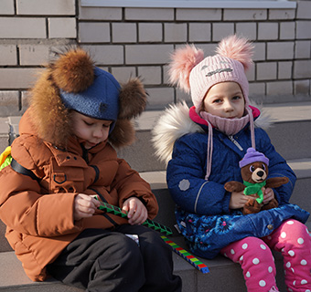 Zwei fünfjährige ukrainische Kinder sitzen in Winterkleidung und mit Rucksäcken auf Treppenstufen.
