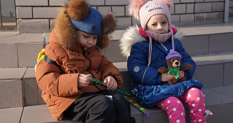 Zwei fünfjährige ukrainische Kinder sitzen in Winterkleidung und mit Rucksäcken auf Treppenstufen.