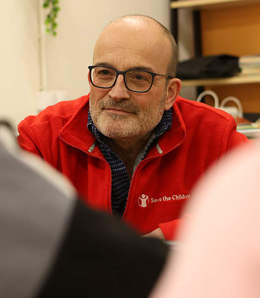 Ein Mann mit Save the Children-Jacke sitzt an einem Tisch und hört zu