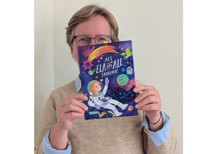 Eine Frau hält ein Buch in die Kamera mit dem Titel: Als Ela das All eroberte