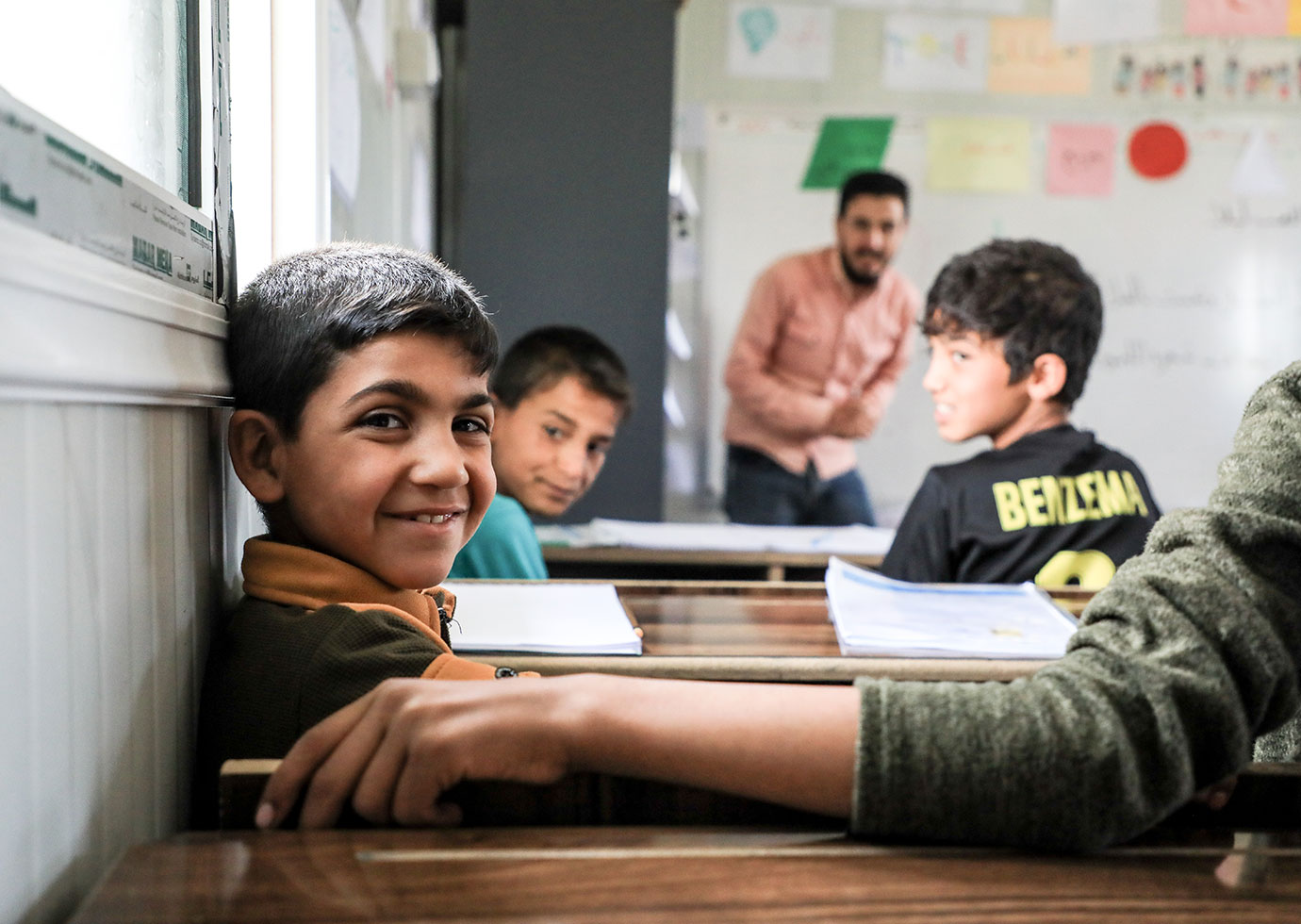 In einem Geflüchtetencamp im Nordosten Syriens kann der achtjährige Waleed* erstmals eine Schule besuchen. © Roni Ahmed / Save the Children Ein Junge in einem Klassenraum lächelt in die Kamera. In der Unschärfe sind mehrere weitere Personen zu erkennen.