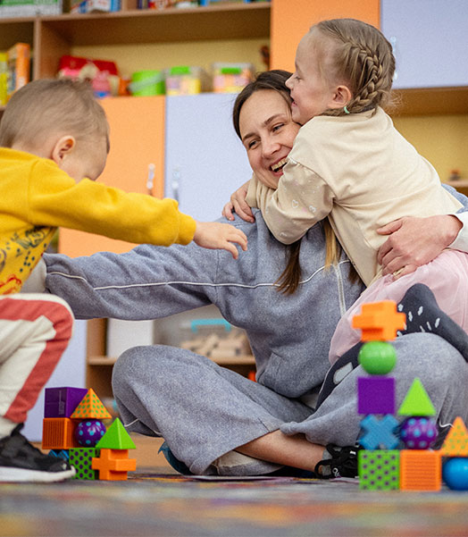Zwei Kinder spielen mit ihrer Mutter in einem Schutz- und Spielraum in der Ukraine. Derartige Projekte kann Save the Children nur dank regelmäßiger Spenden umsetzen. © Danil Shevchenko / Save the Children Eine lächelnde Frau, die auf dem Boden sitzt und zwei Kinder in ihren Armen hält.
