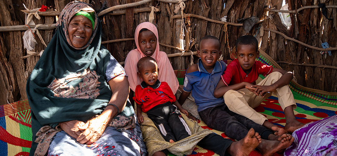Abdi mit seiner Familie © Mustafa Saeed / Save the Children Eine erwachsene Person und vier Kinder sitzen auf dem Boden und lächeln in die Kamera.