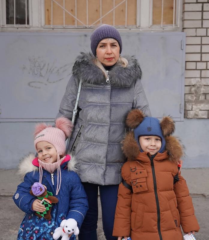 Eine Mutter steht mit ihren zwei Kindern in Winterkleidung vor einer Hauswand.