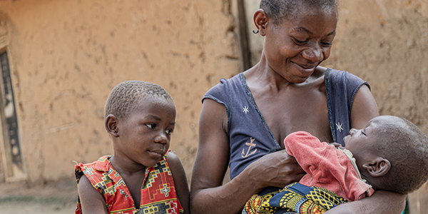 Mutter Charly* hält Gervienne*, 15 Monate, im Arm, die von Save the Children Spezialnahrung gegen Mangelernährung erhalten hat. © Patou Dombi/Save the Children Eine Mutter sitzt mit ihrem Kleinkind im Arm und lächelt.
