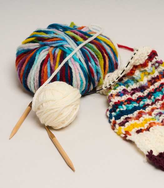 Die Aktion „Ein Schal fürs Leben“ von Brigitte und Save the Children lädt auch 2026 zum Stricken für den guten Zweck ein. © Save the Children Zwei Wollknäuel, der Anfang eines gestrickten Schals und Stricknadeln liegen auf einem Tisch.