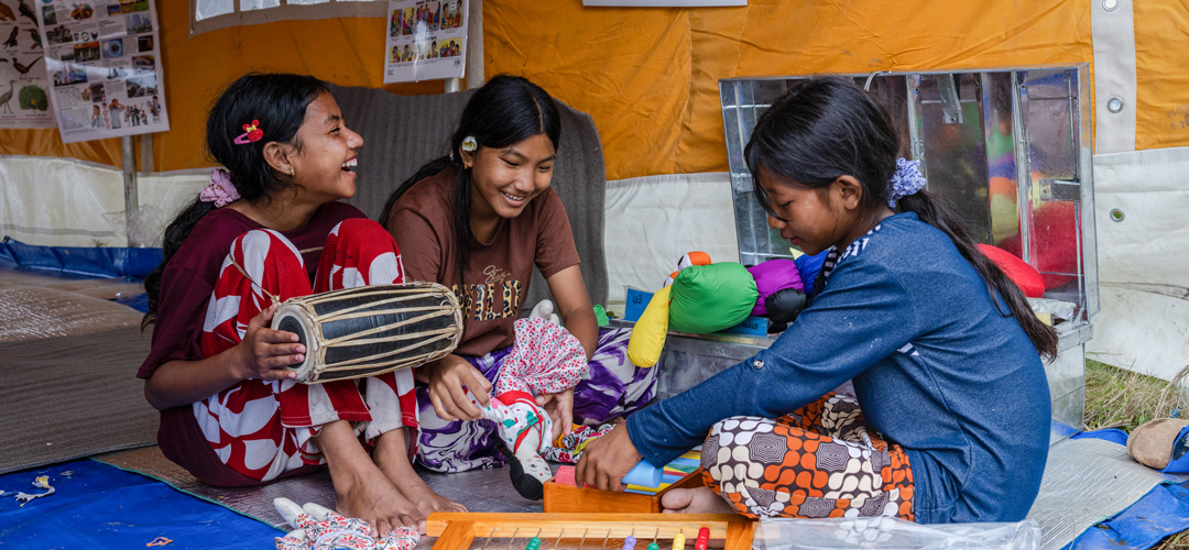 Sapana* und Kala* (links im Bild) rannten jeweils allein bergauf, als das Wasser ins Tal schoss. Unser Schutz- und Spielraum hilft ihnen, das Erlebte zu verarbeiten. © Ulet Ifansasti / Save the Children Drei Mädchen sitzen zusammen und lachen. Um sie herum liegt Spielzeug.