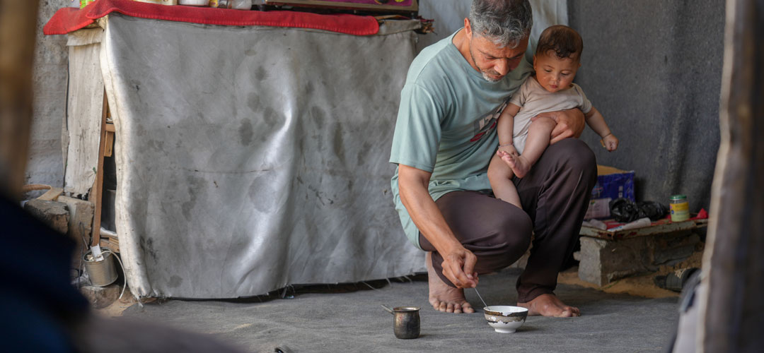 Kleidung für die Kinder, einen Gaskocher, Windeln für Sana* und Haushaltsgegenstände: Dafür braucht Raheem* die Bargeldhilfen von Save the Children. © Save the Children Ein Mann kniet auf dem Boden und füttert ein Kind auf seinem Arm. Hinter ihnen sieht man Zeltplanen.