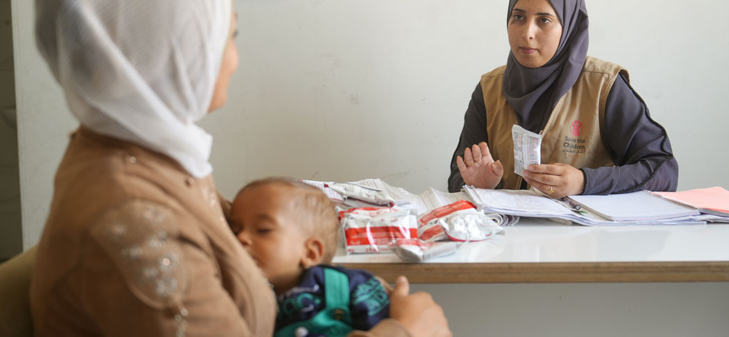 Schwanger im Krieg: Yasmin* hatte kaum genug Essen, um selbst zu überleben. So kam ihr Sohn krank zur Welt. Unser Team half ihnen unter anderem mit Spezialnahrung. © Save the Children Eine Frau mit einem Kind auf dem Arm sitzt gegenüber einer Save the Children-Mitarbeiterin. Sie sprechen miteinander.