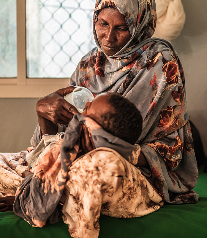 Somalia: Infolge einer Dürre wurde Ismail (Name geändert) mangelernährt. In unserem Ernährungszentrum fand seine Mutter Hilfe. Hier kommt er wieder zu Kräften. © Brian Otieno / Save the Children Eine Frau stützt ihr Kind, das in ihrem Schoß liegt. Sie gibt ihm etwas zu trinken aus einem Becher.