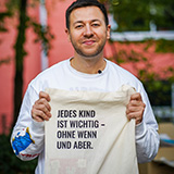 Portrait eines Mannes mit kurzen braunen Haaren, der eine Tasche hoch hält.