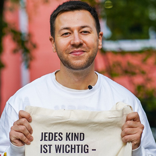 Portrait von eines Mannes mit kurzen braunen Haaren, der eine Tasche hoch hält.